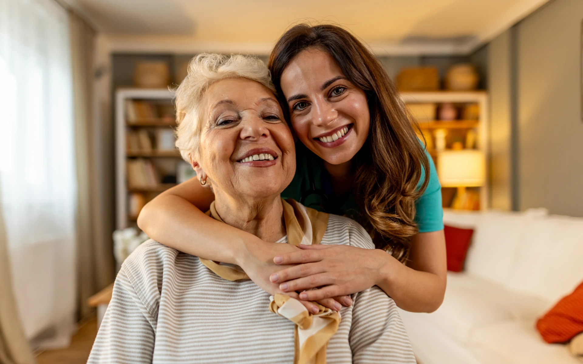Smiling woman hugging elderly lady at home.